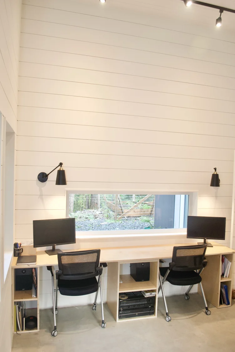 Modern two-story home with large glass windows, warmly lit interiors, and a balcony. Surrounded by trees and a manicured lawn at dusk. Cozy ambiance.