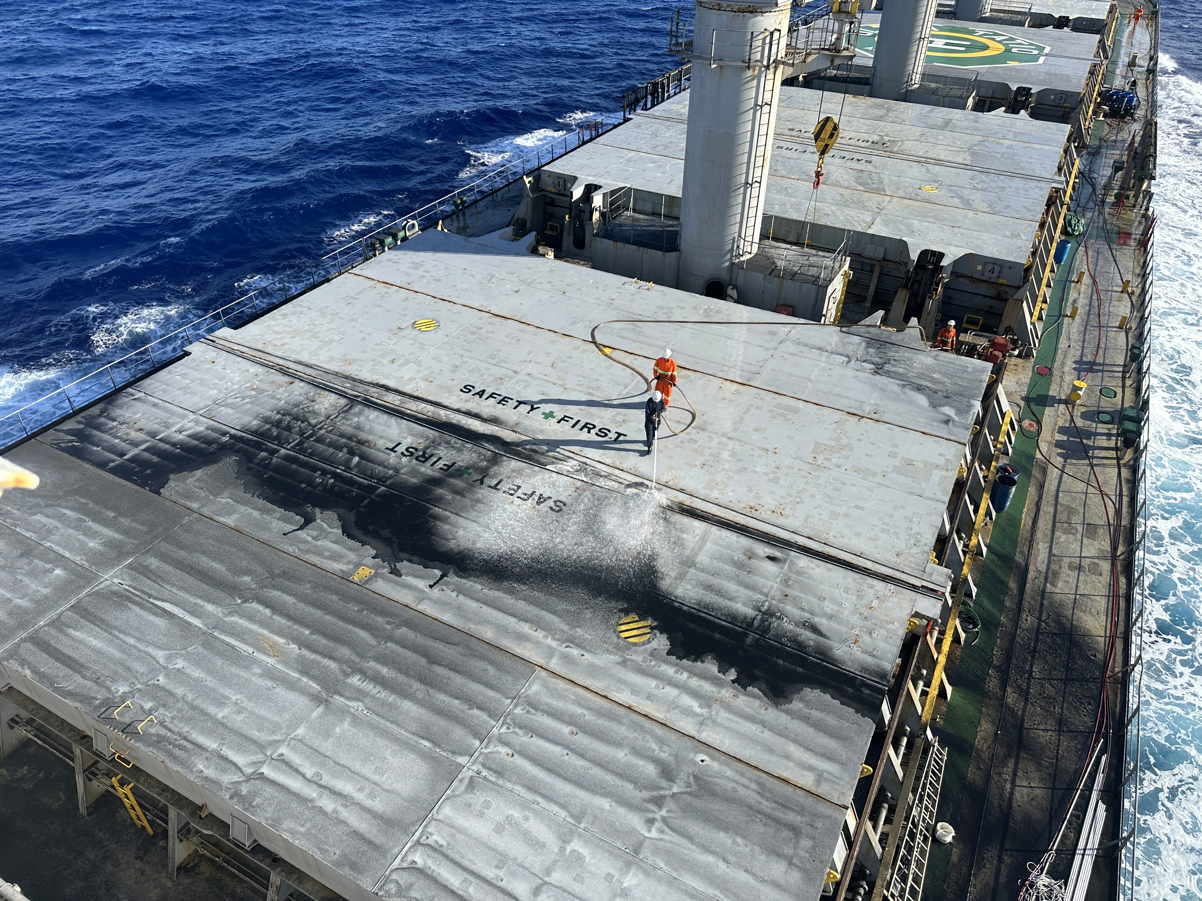 Η ομάδα Seachios® Marine Services προβαίνει στον καθαρισμό των καλυμμάτων θυρών καταστρώματος με χρήση υψηλής πίεσης στη θάλασσα κατά τη διάρκεια του ταξιδιού από το Καμπεδέλλο προς το Σάντος, προετοιμάζοντας τις αποθήκες φορτίου να ανταποκρίνονται στα αυστηρά πρότυπα καθαριότητας, ενώ το φορτηγό πλοίο πλέει στα ανοιχτά της βραζιλιάνικης ακτής, με χαρακτηριστικές επισημάνσεις “ΑΣΦΑΛΕΙΑ ΠΡΩΤΑ” και πλήρη συμμόρφωση με τα μέσα ατομικής προστασίας.