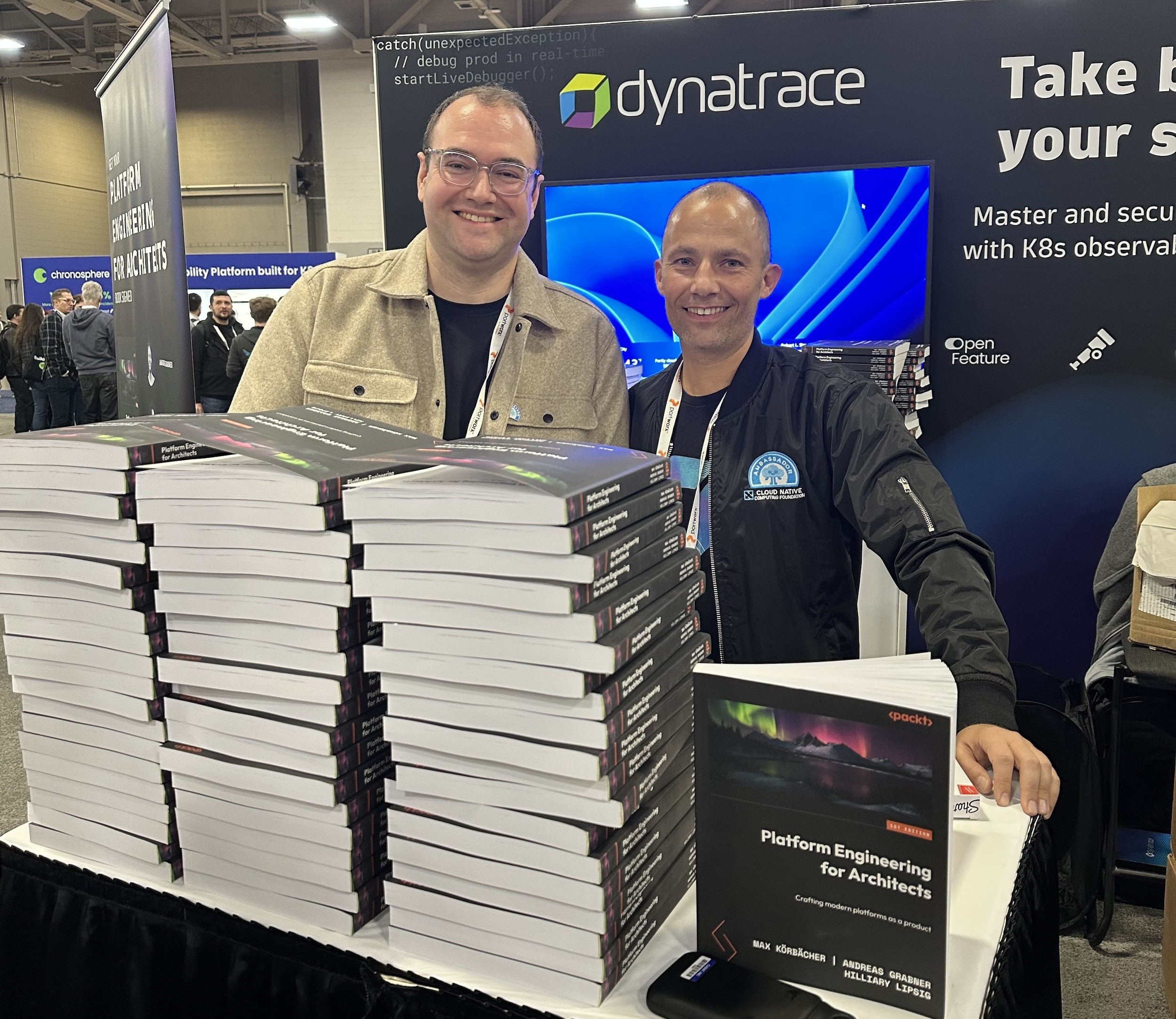 Zwei Autoren hinter einem großen Stapel des Buchs Platform Engineering for Architects am Dynatrace-Stand während der Buchsignierstunde auf der KubeCon