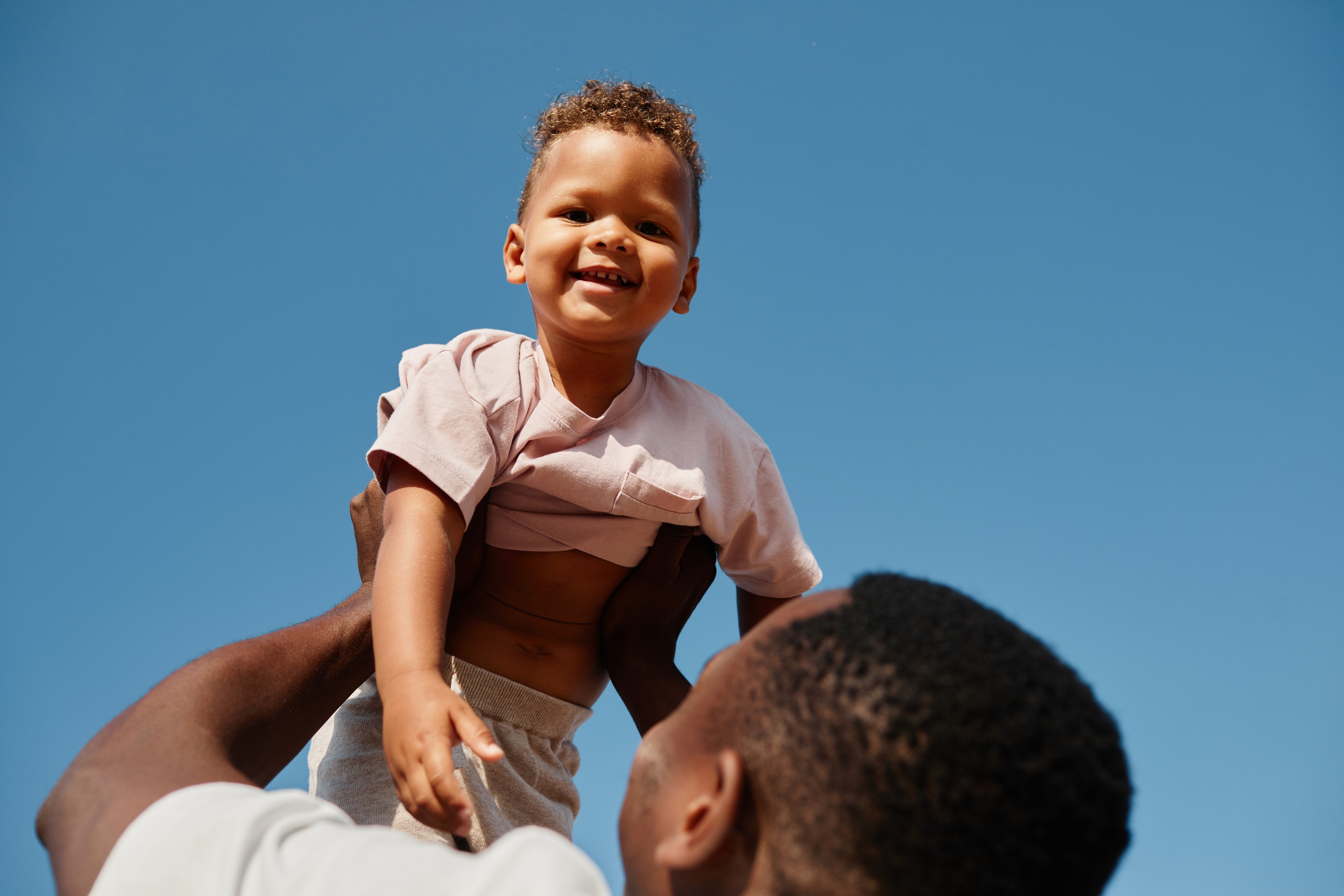 Smiling toddler being lifted into the air by an adult against a clear blue sky.