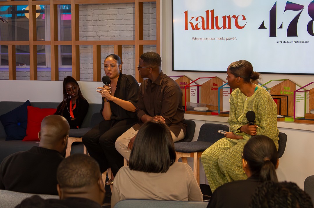 A young person with curly hair smiles at the camera while sitting in front of a laptop against a dark background.