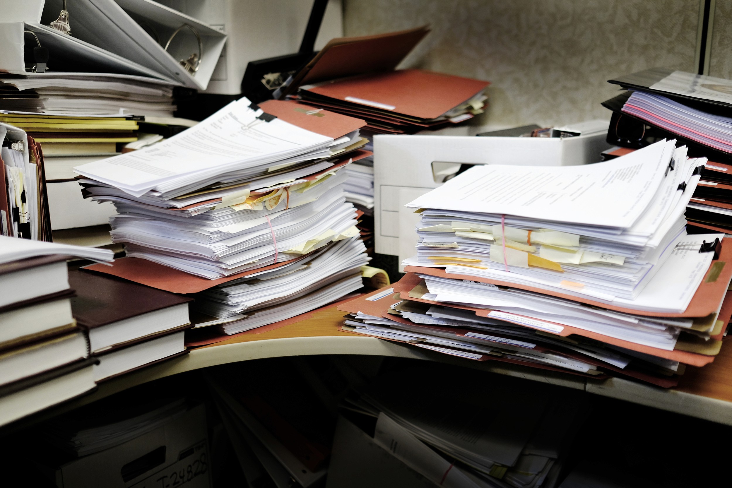 Detail of a messy desk with books binders files and papers stacked up high in a disorganized mess