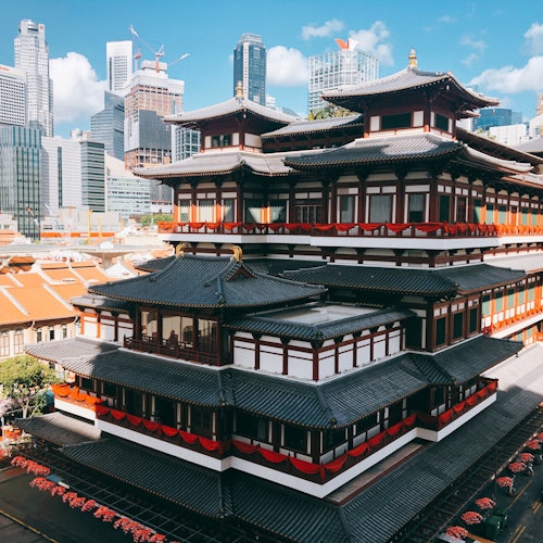 一座带有红色装饰的多层传统建筑，坐落在蓝天下现代摩天大楼的背景中。