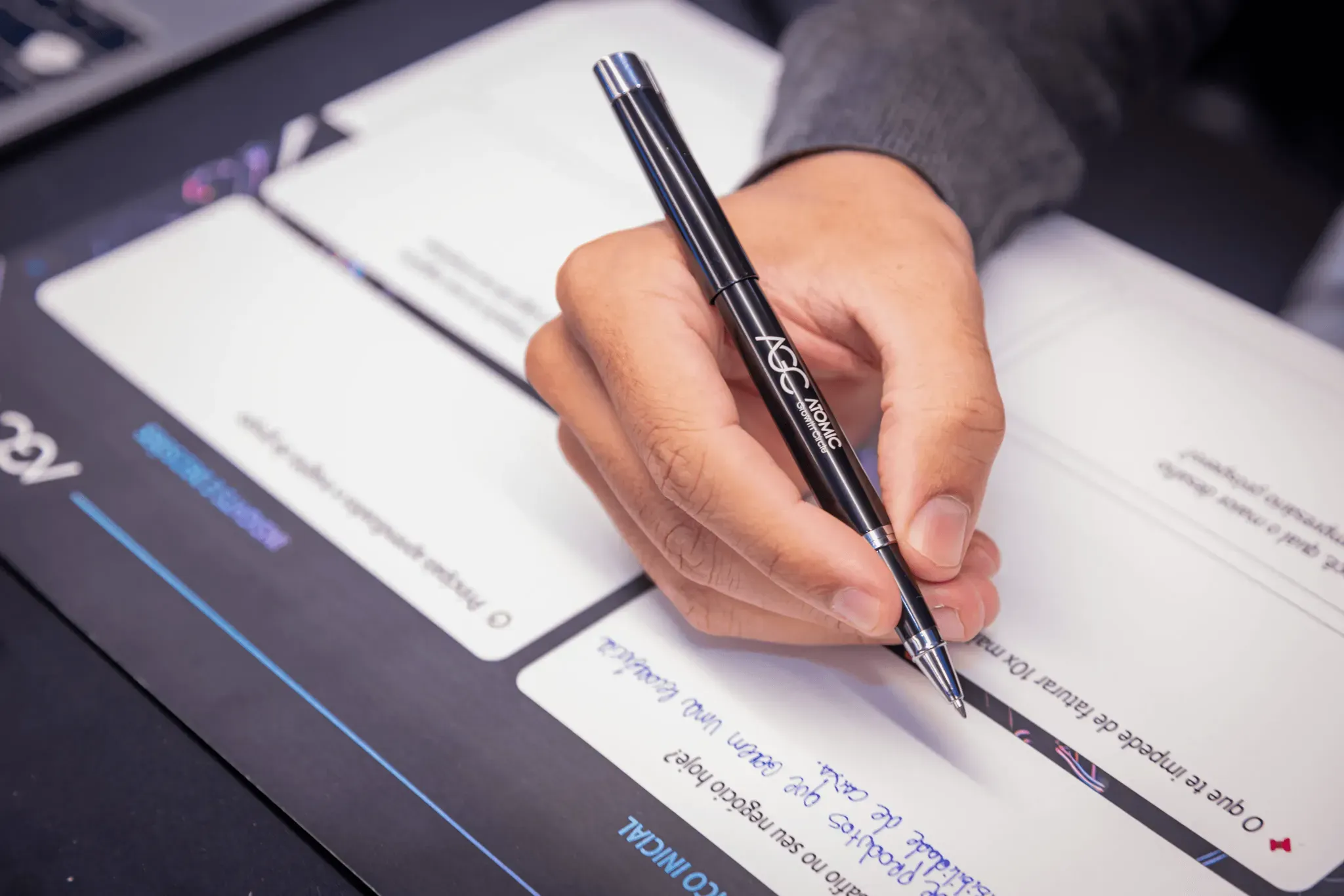 A hand holding a pen, signing a document on a clipboard. Atomic Growth Club. Atomic Group. Br24. 