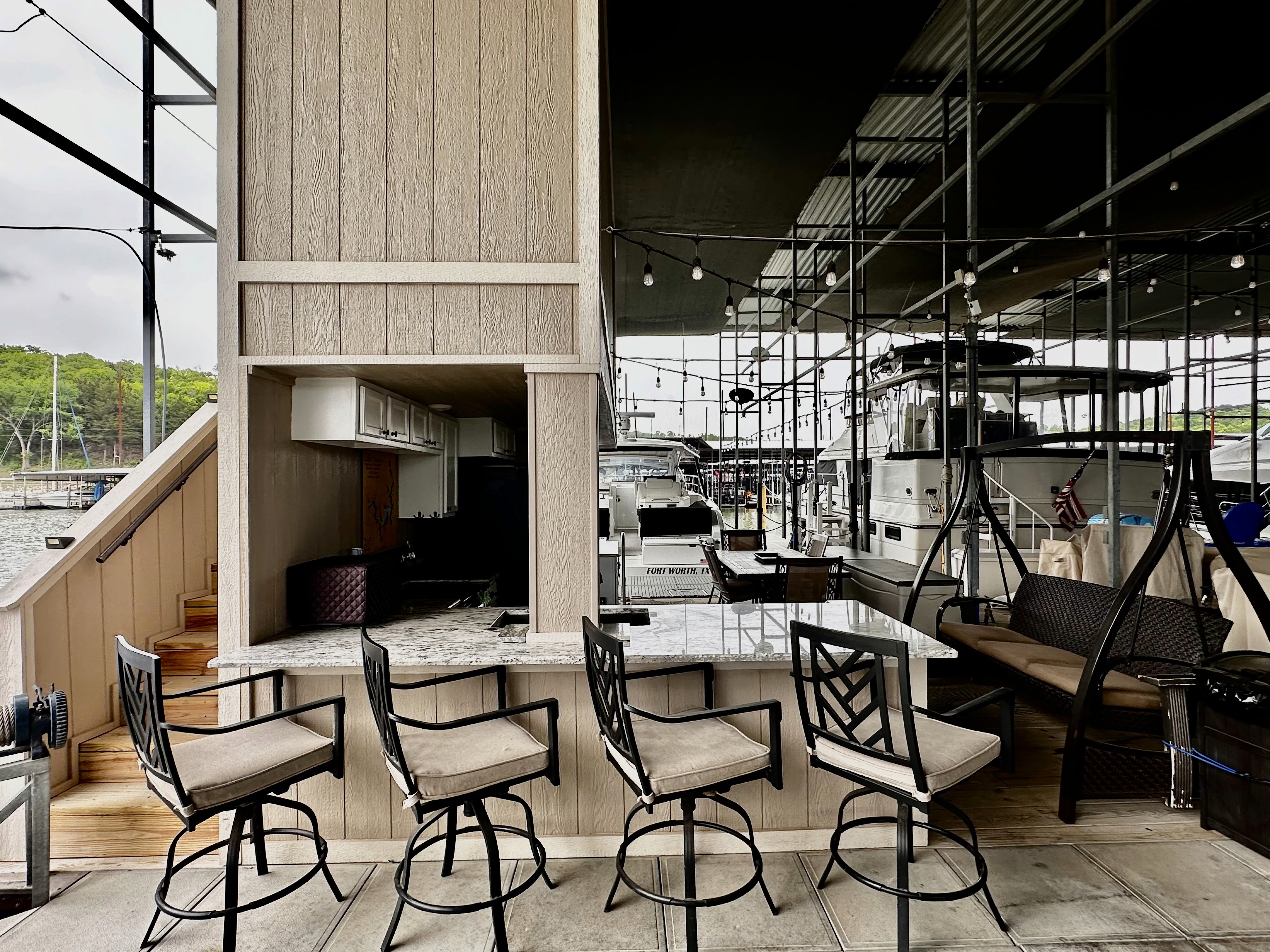 A nautical-themed outdoor bar area on a dock features a granite countertop, four cushioned swivel chairs, and reflections of nearby boats on the polished wood surfaces, creating a relaxed marina atmosphere.