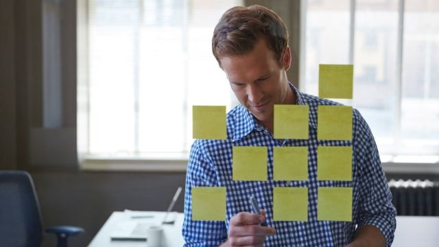 Homem branco de camisa xadrez azul observando post-its organizados em parede de vidro à sua frente em ambiente de escritório