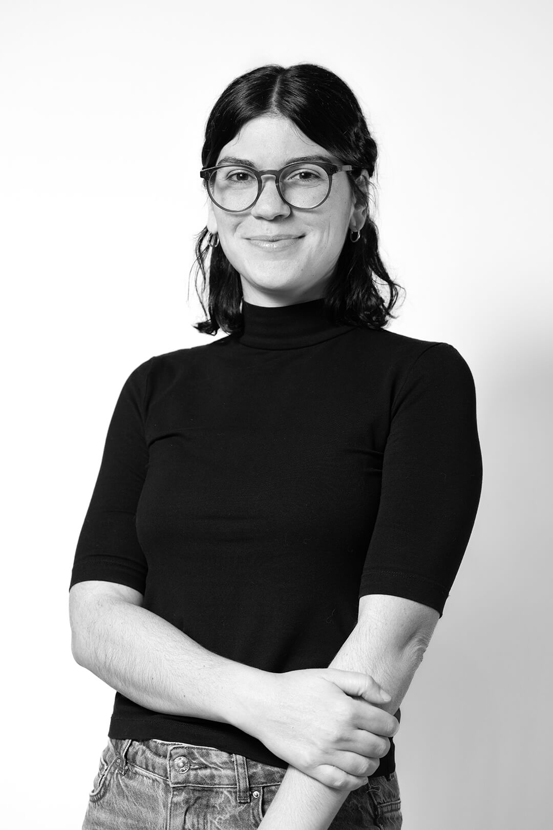 A person in glasses and a black turtleneck smiles confidently at the camera. Their arms are crossed, conveying a friendly, approachable demeanor.