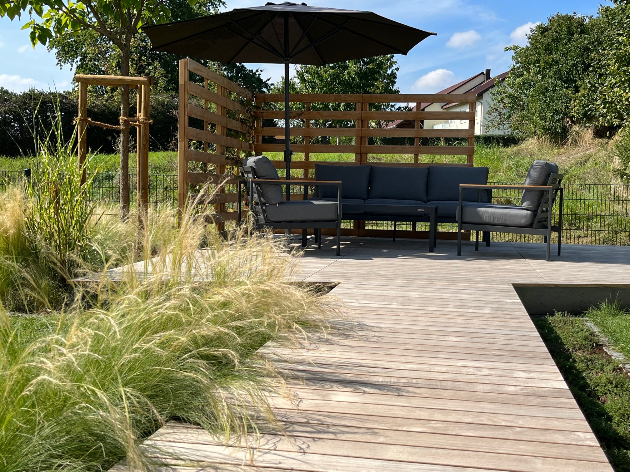 Gartenbau von Marohn & Binder mit Holzterrasse, Möbeln und Sonnenschirm vor einem Holzzaun.