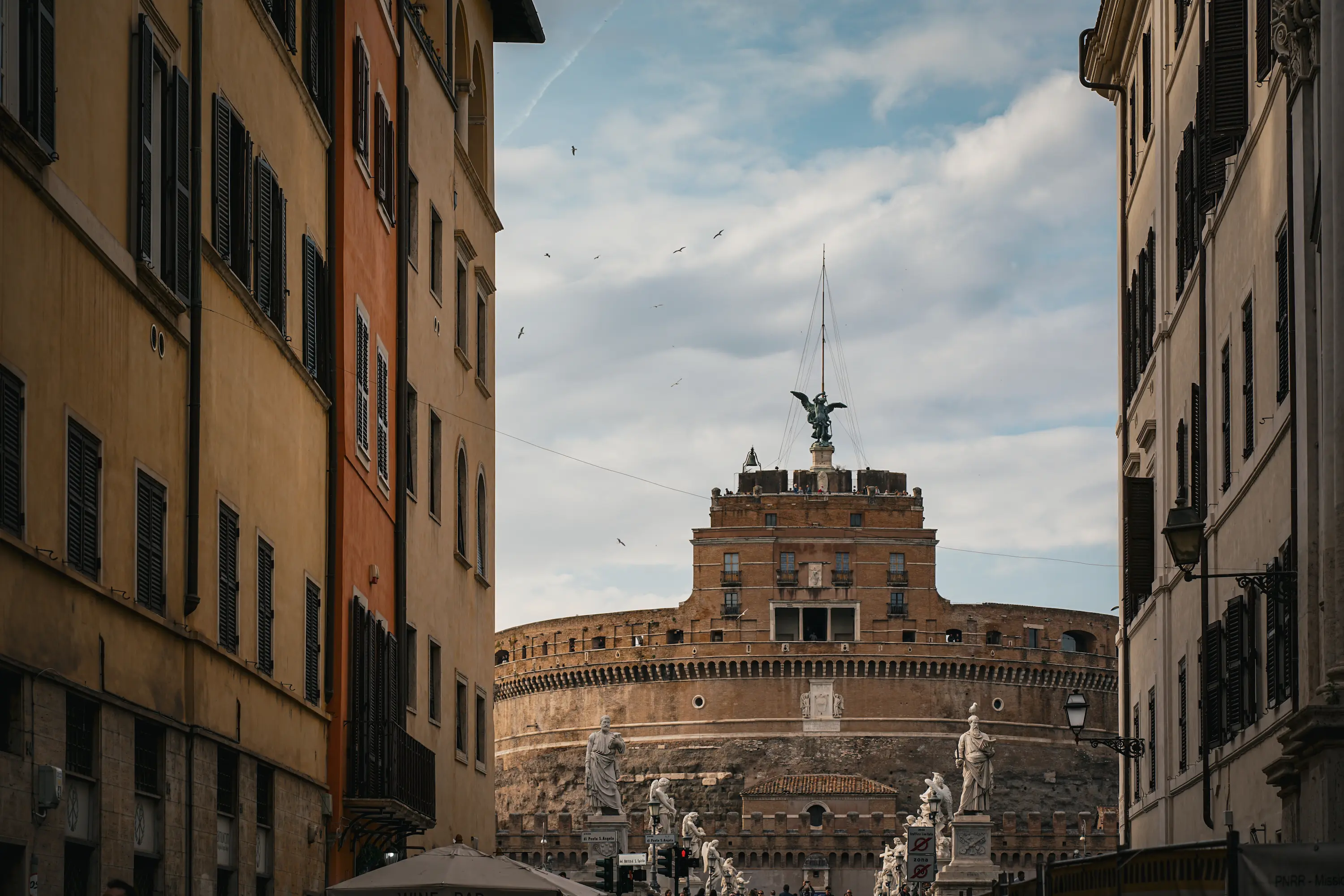 Vue du Castel Sant’Angelo à Rome, encadré par les façades d’une rue, sous un ciel nuageux avec plusieurs oiseaux en vol