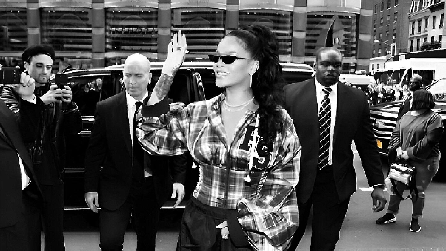 Black and white street photo with a multi-stories carpark on background. On close-up is a celebrity that just dropped of the limo, waving at  her fans and media. She is protected by 2 executive protection officers. 