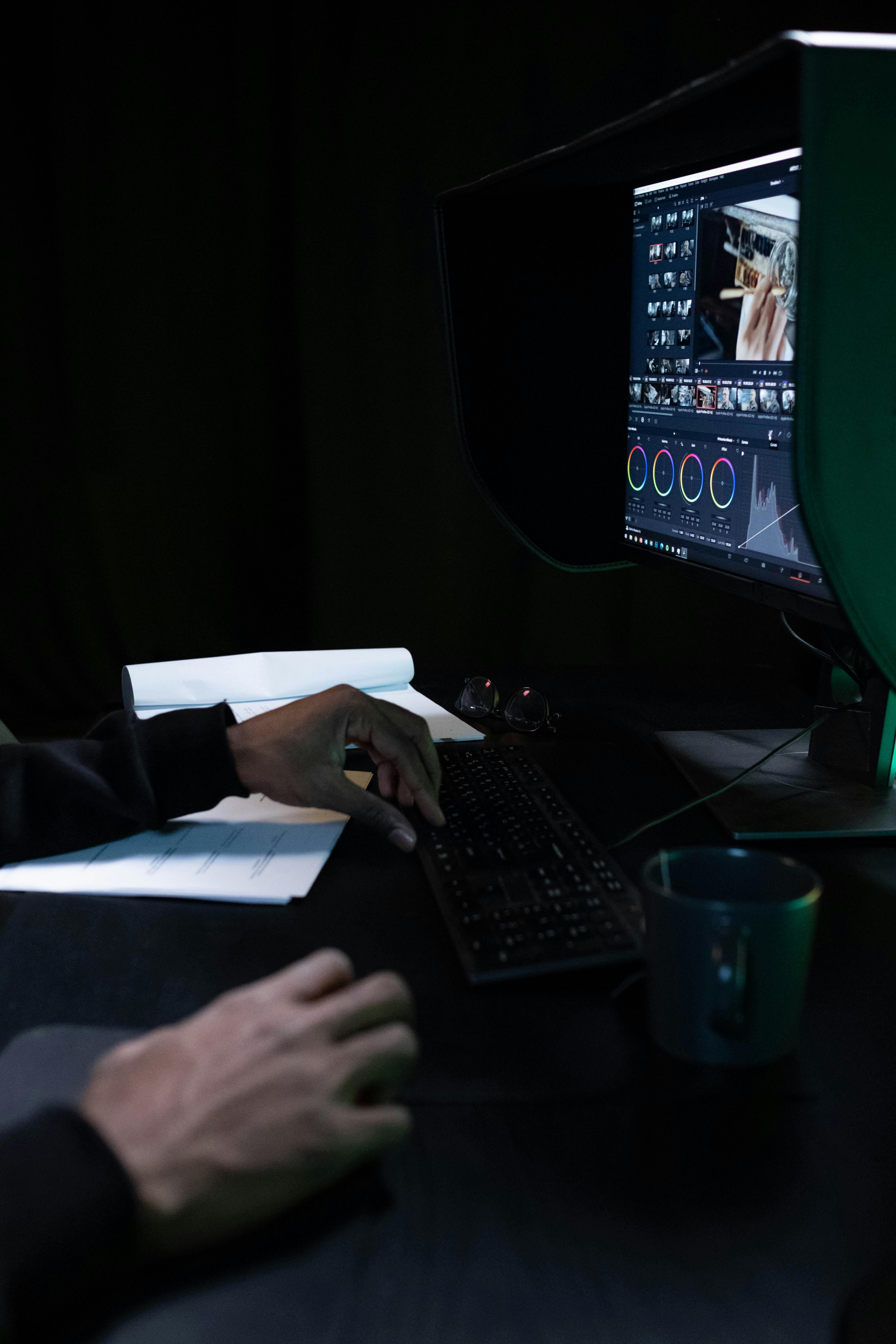 Hands color grading video on a hooded monitor in a dark room. A keyboard, papers, and a mug sit on the desk.