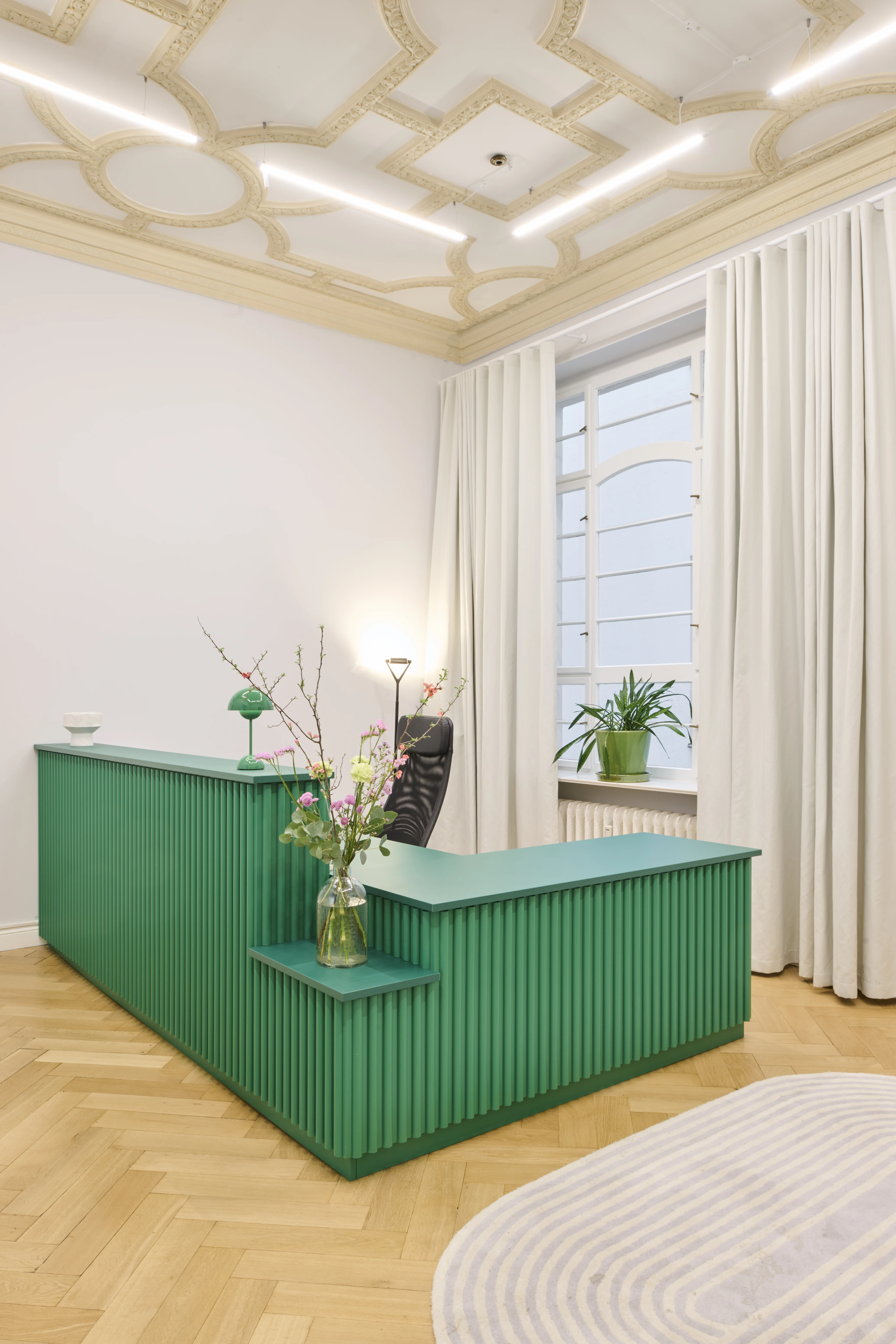 Reception at INTERMATE BERLIN with an emerald-green slatted stepped counter, ornate Art Nouveau ceiling with integrated light lines, cream curtains and herringbone parquet.