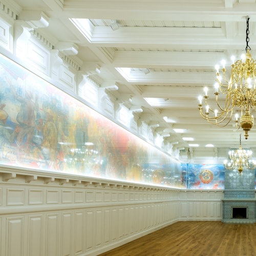 Elegáns terem magas mennyezettel, csillárokkal, a falon hosszú freskóval, fehér lambériával és fa padlóval. Nincs jelenlévő ember.