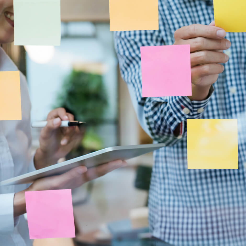 Two people brainstorming with colorful sticky notes on a glass wall; one holds a tablet and pen, the other places a pink sticky note.