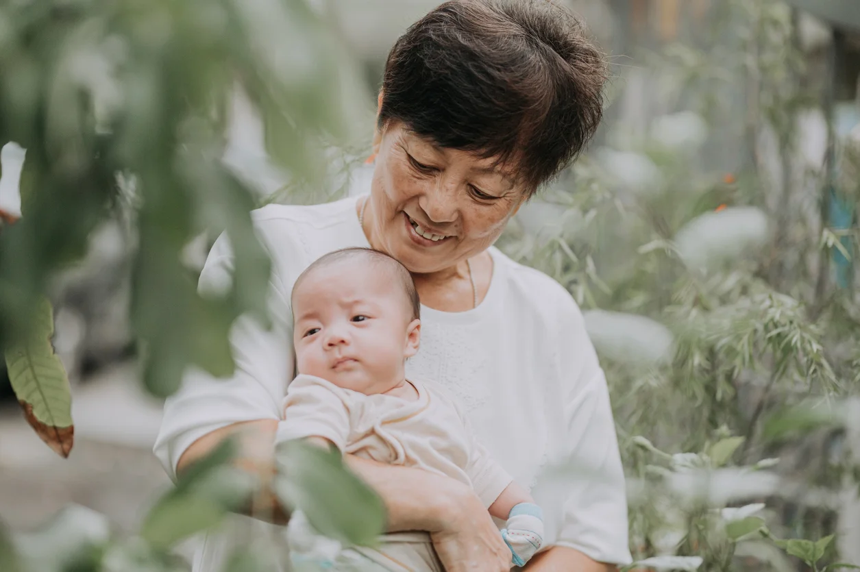 Elderly nanny lovingly holding a baby outdoors, symbolising trusted confinement care in Singapore.