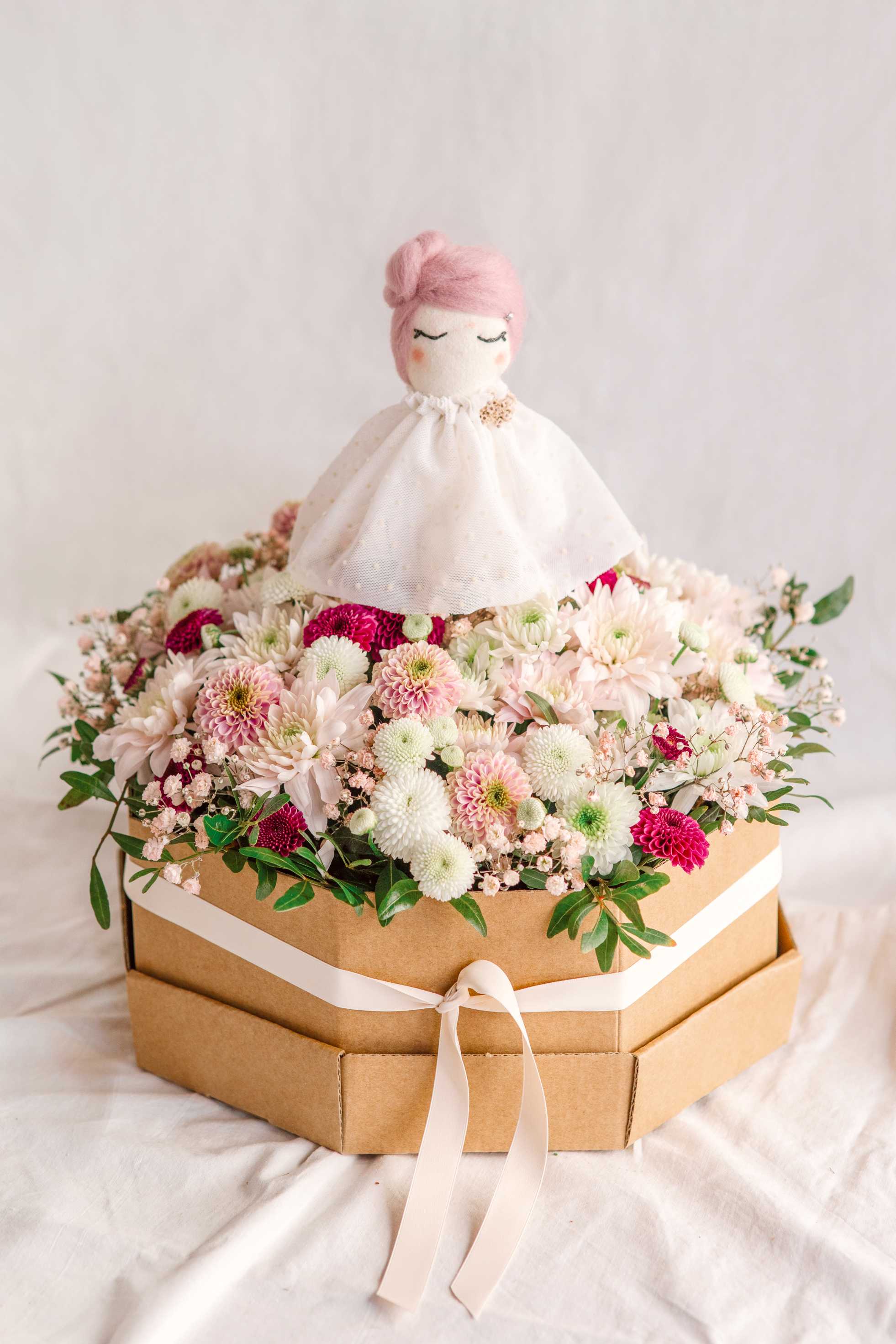 Arranjament de flors silvestres en tons suaus dins una caixa octogonal de cartró kraft amb cinta blanca, amb una nina decorativa al centre.