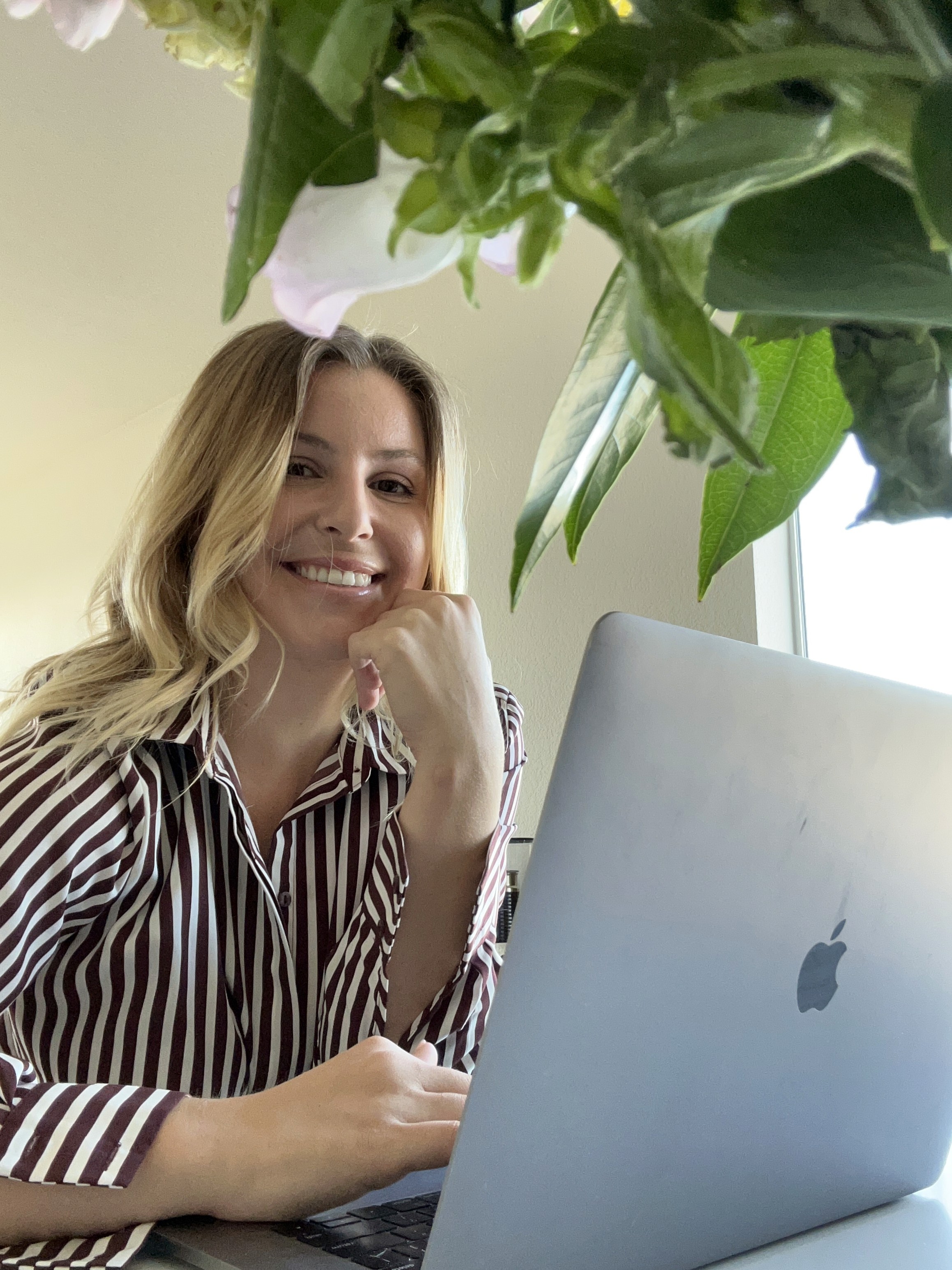 A smiling man with curly hair is sitting at a desk, looking at the camera, with a laptop and a lamp in the background.