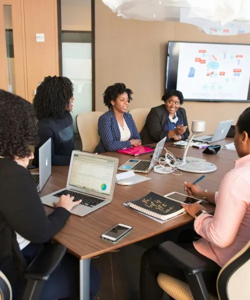 A diverse group of businesswomen in a meeting around a table, collaborating with laptops and tablets.