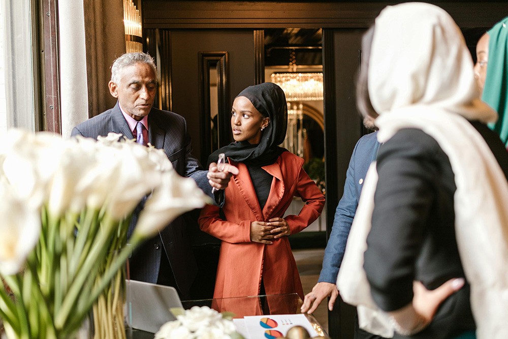 A professional woman wearing a hijab and rust-colored blazer gestures while speaking during a business meeting in an elegant office setting. She stands at a conference table with colleagues, including an older man in a suit and others in business attire. White flowers and documents are visible on the table, illustrating diverse representation and active participation in corporate leadership.