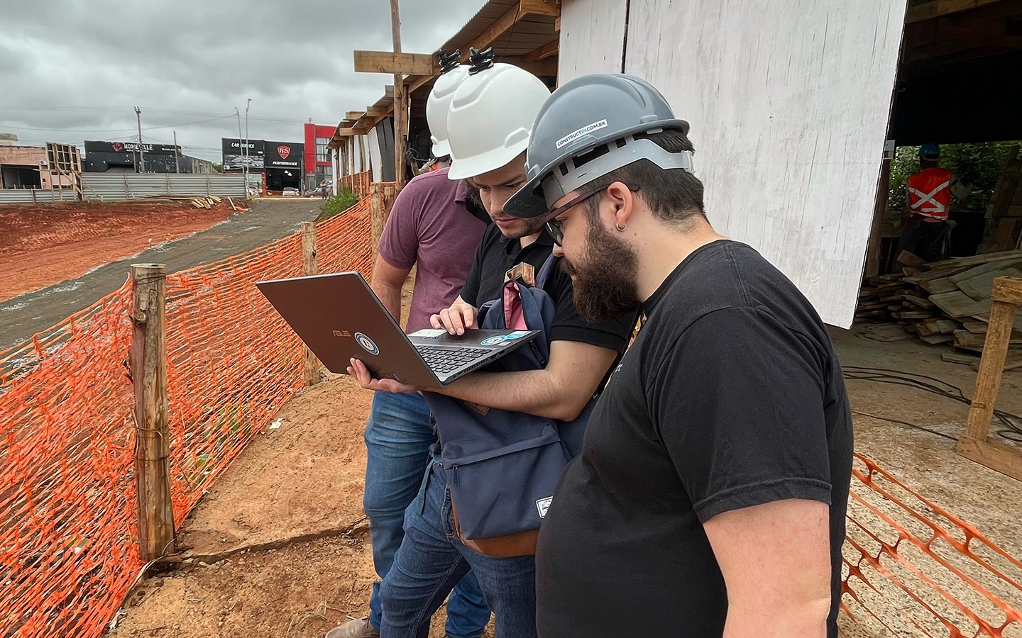 Três colaboradores da Construct IN usando capacetes de segurança (um cinza com o logo da empresa e dois brancos) em um canteiro de obras. Eles estão reunidos em torno de um notebook, analisando dados de Captura da Realidade diretamente no campo, promovendo uma gestão transparente.