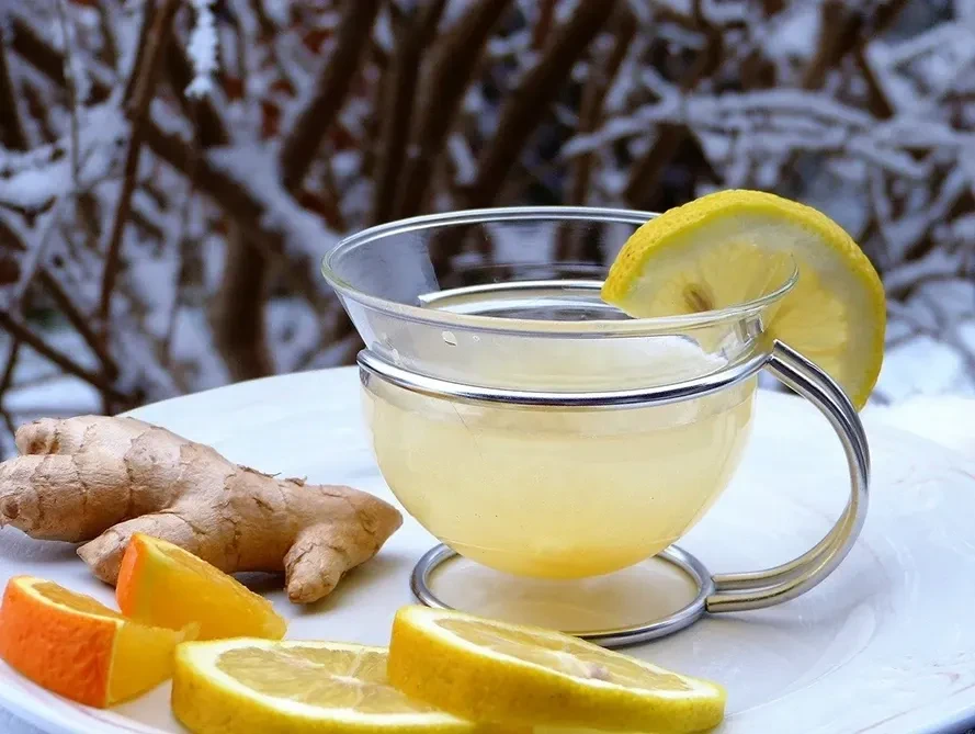 Clear glass cup of lemon ginger tea with a lemon slice on the rim, surrounded by fresh ginger and citrus slices on a snowy backdrop. Cozy and warming vibe.
