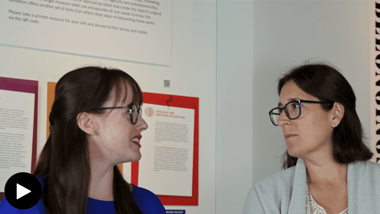 Carlee Forbes and Erica Jones looking at each other with wall text from the Belongings exhibition in the Belongings exhibition