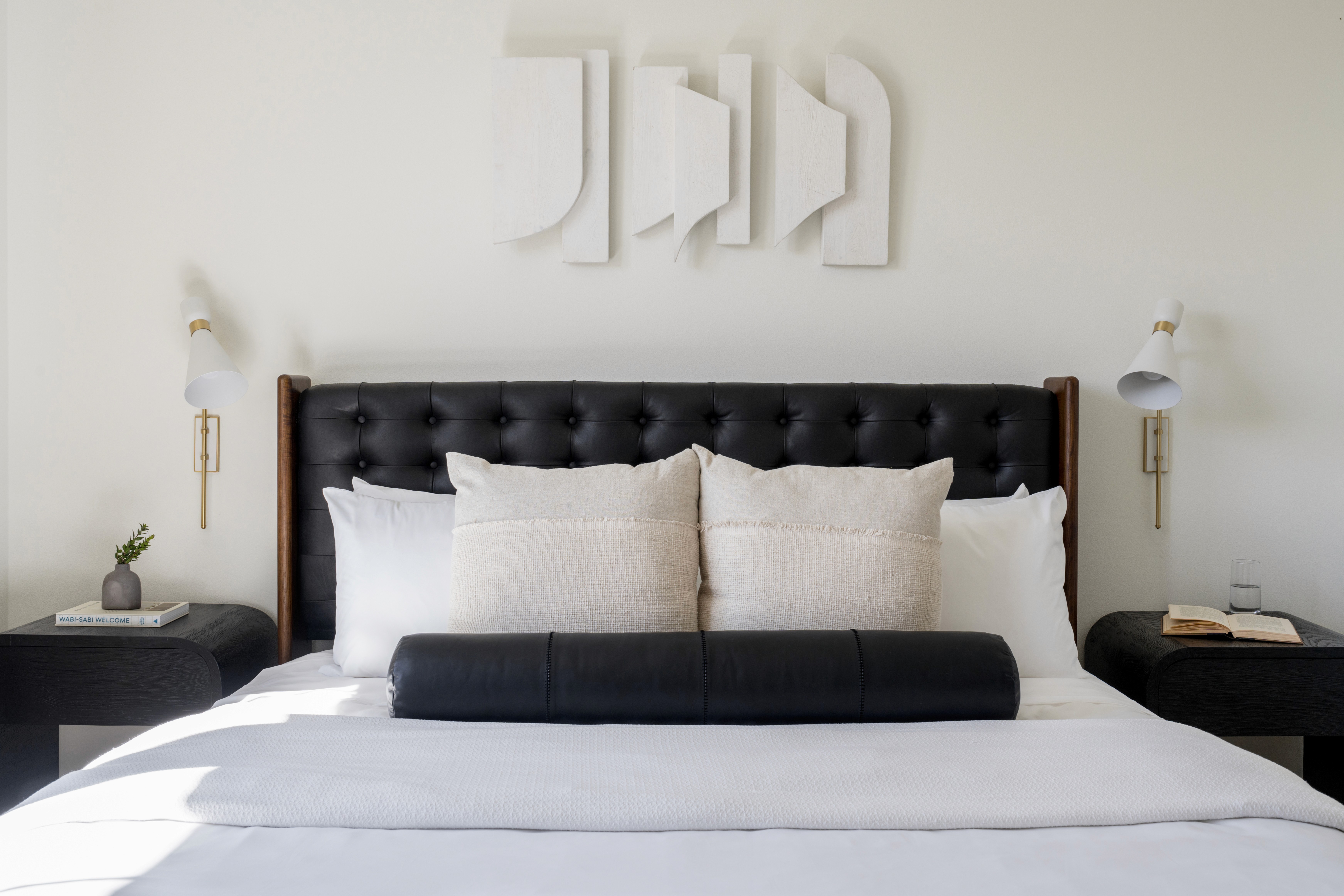 Dark wood and black faux leather upholstered bed frame, white and cream bedding, black leather lumbar pillow, sconce lighting, black stained wood side tables with rounded edges, and white washed wood geometrical art piece above bed