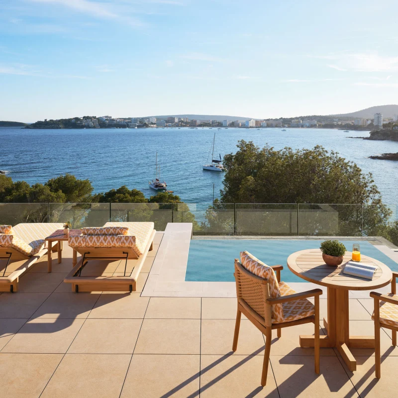 Ein Blick aus der Luft auf ein luxuriöses Küstenresort mit einem großen Pool, das auf einer felsigen Halbinsel liegt, mit einer Stadt und Bergen im Hintergrund.