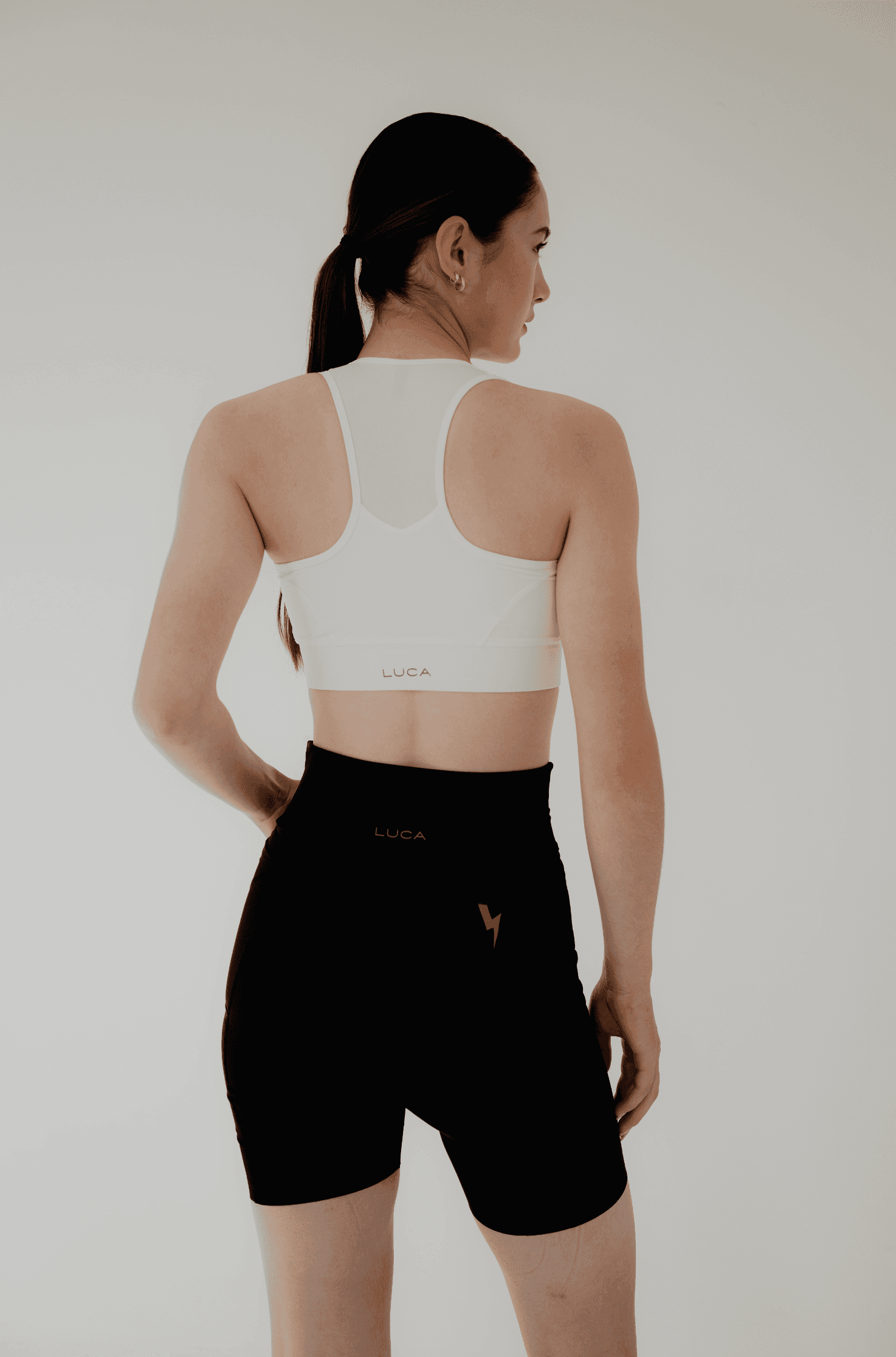 Back-view-of-a-woman-modeling-a-white-spots-bra-with-mesh-panel-and-LUCA-logo-paired-printed-with-black-high-waisted-biker-shorts-featuring-a-reflective-lighting-bolt-graphic