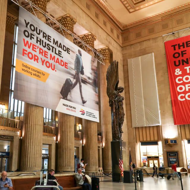A large banner advertising online courses for working adults hangs in a grand train station with people sitting and a statue visible below.