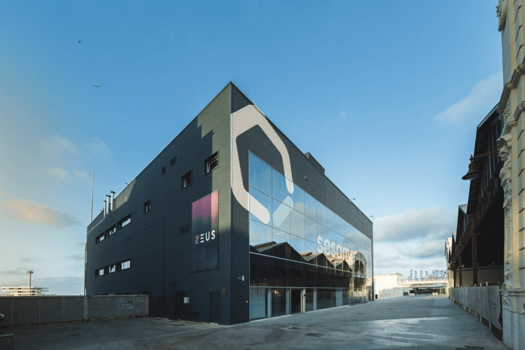 Modern industrial-style building with dark exterior panels, large glass windows, and visible logos for ZEUS and Sesame.