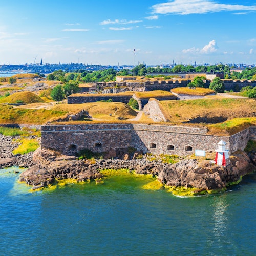 Ιστορικό φρούριο με πέτρινους τοίχους, περιτριγυρισμένο από νερό και πράσινο, κάτω από έναν γαλανό ουρανό με διάσπαρτα σύννεφα.