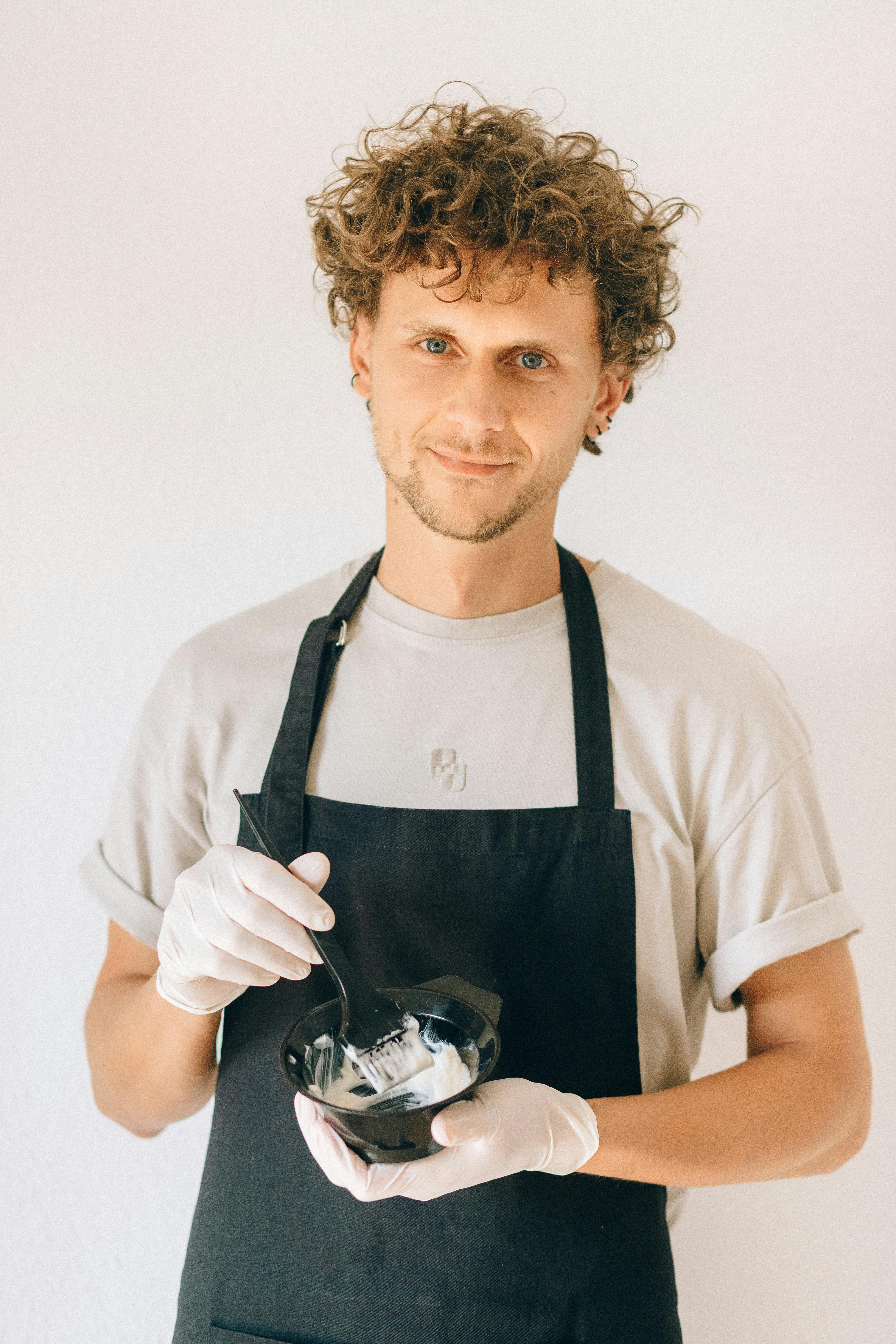Man in apron mixing hair dye with a smile, wearing gloves, holding a bowl with dye and a mixing stick.