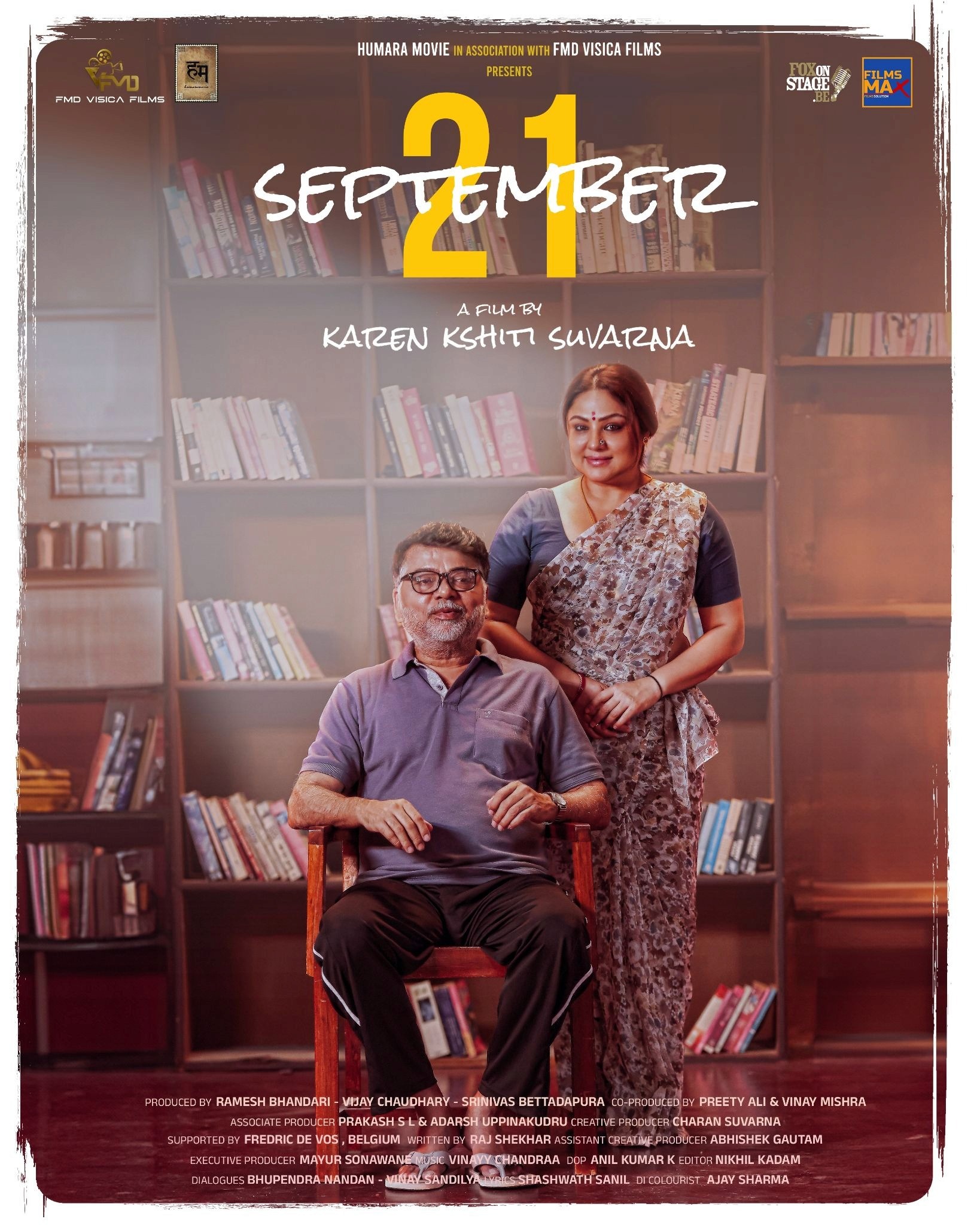 A man seated in a library, smiling, with a woman standing behind him. The text "September 21" is above in bold. Bookshelves form the backdrop.