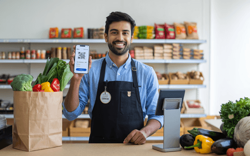 Smiling cashier at a grocery store counter holding a smartphone displaying a QR code powered by Razorpay for digital payment beside a bag of fresh vegetables.