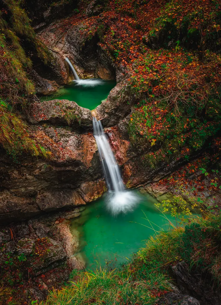A serene waterfall cascades into a vibrant green pool, surrounded by rocky cliffs and autumn foliage in Soča Valley, Slovenia.
