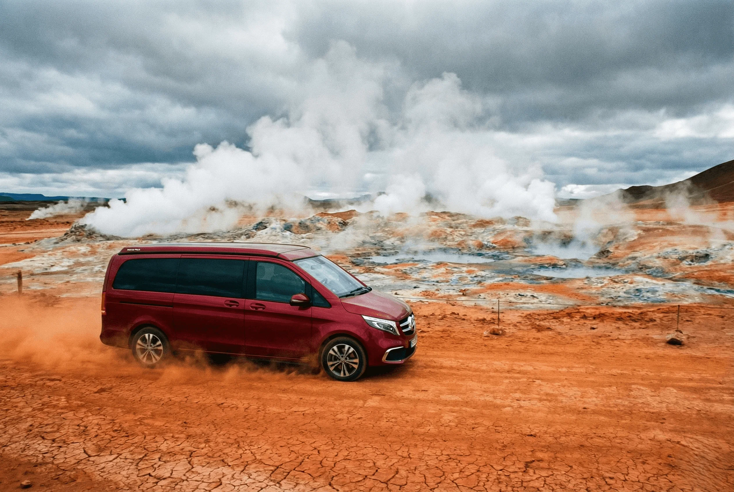 red camper van driving on a dirt road through a geothermal area with steam rising from the ground.