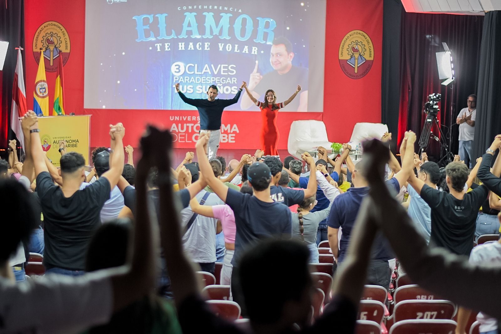 Jorge Baena en el escenario junto a una presentadora durante la conferencia “El amor te hace volar”, frente a un auditorio lleno de asistentes con los brazos levantados en señal de entusiasmo en la Universidad Autónoma del Caribe.