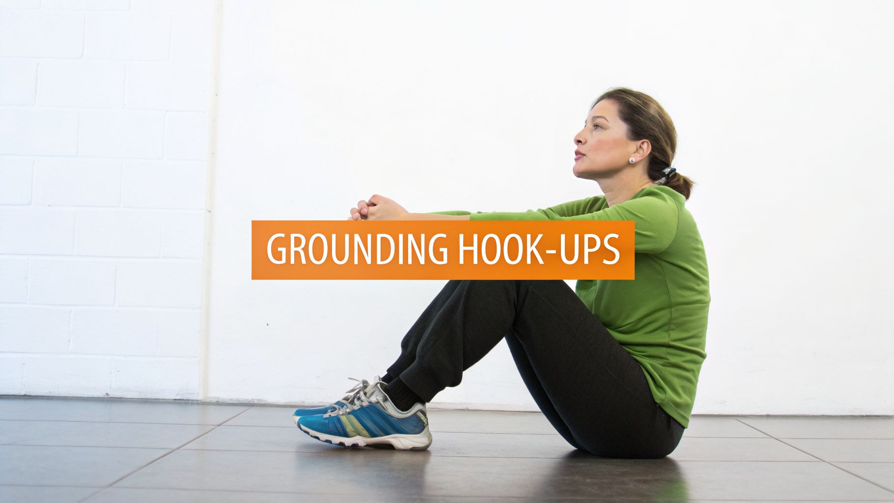 A woman sits on the floor with bent knees, hands clasped, demonstrating a grounding hook-ups exercise.