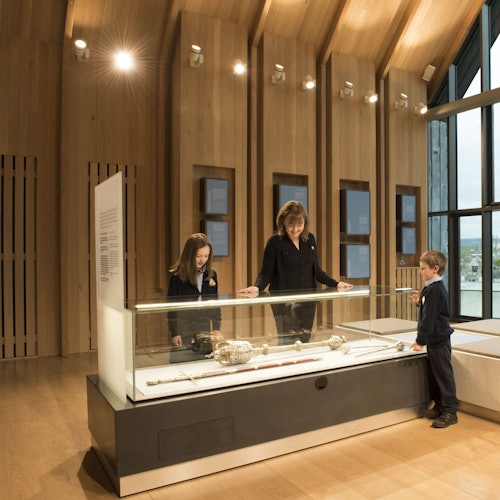 三个人在一个现代化的木饰房间内，通过大窗户在博物馆展示柜中仔细观察文物。