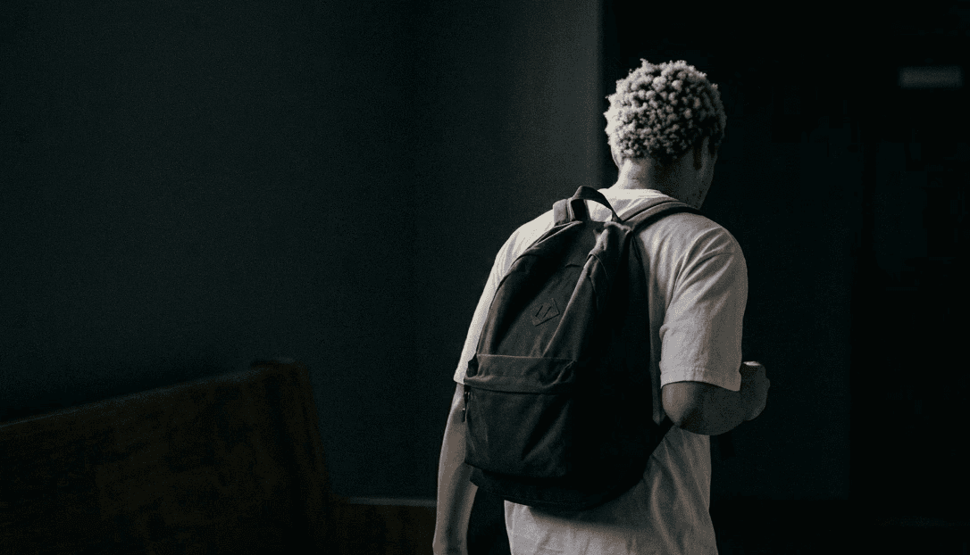 A person with curly hair walks down a dimly lit hallway wearing a casual t-shirt and a backpack, creating a mysterious and introspective atmosphere.
