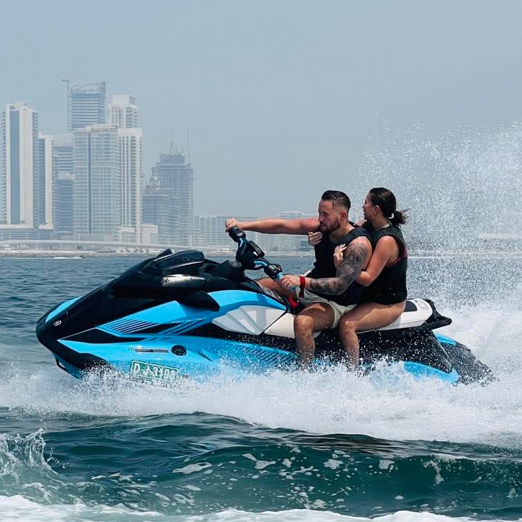 Two people riding on a blue jet ski with a view of the Dubai city skyline in the background.
