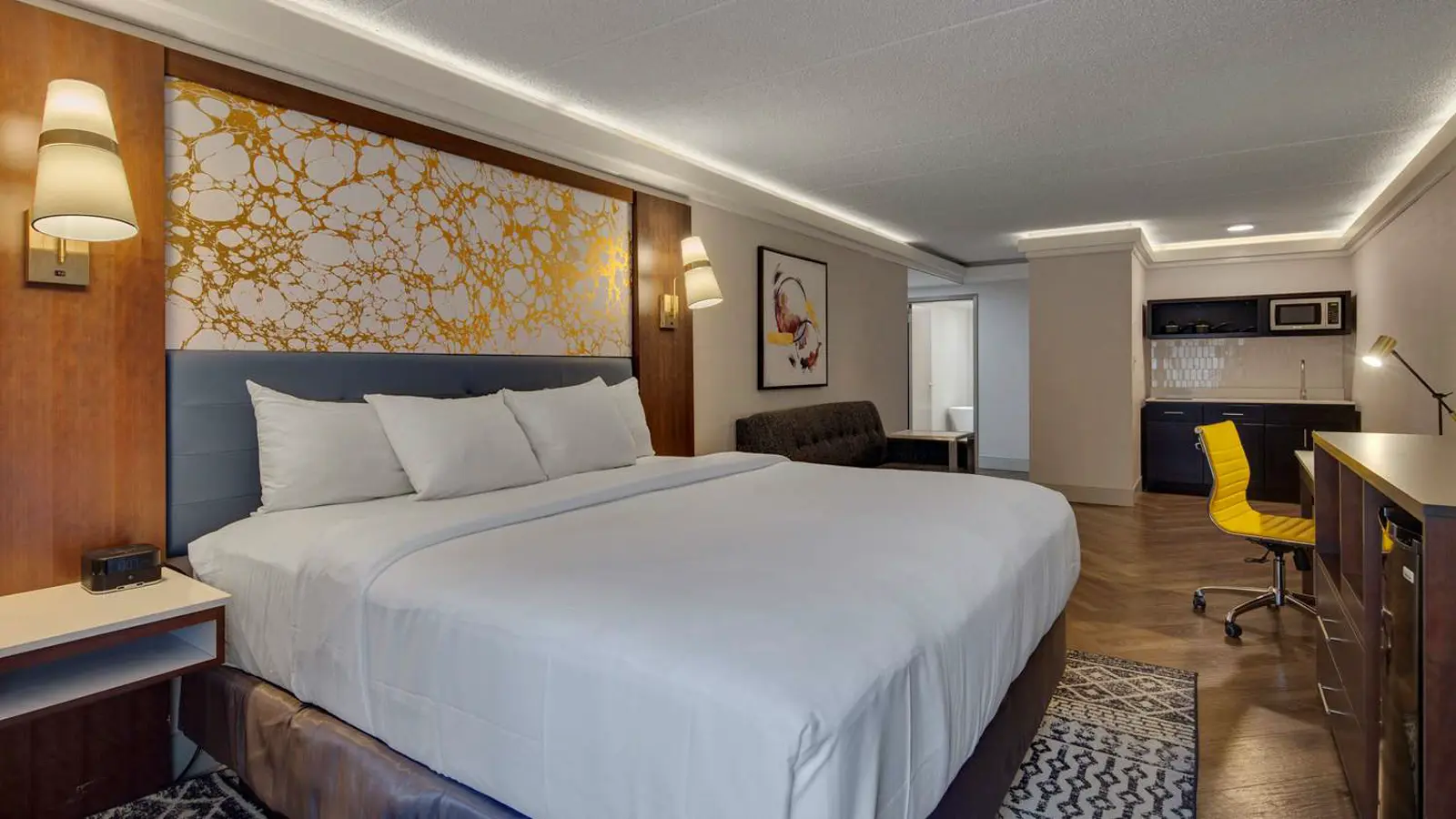 Modern hotel room with king bed, kitchenette, and bright yellow desk chair.