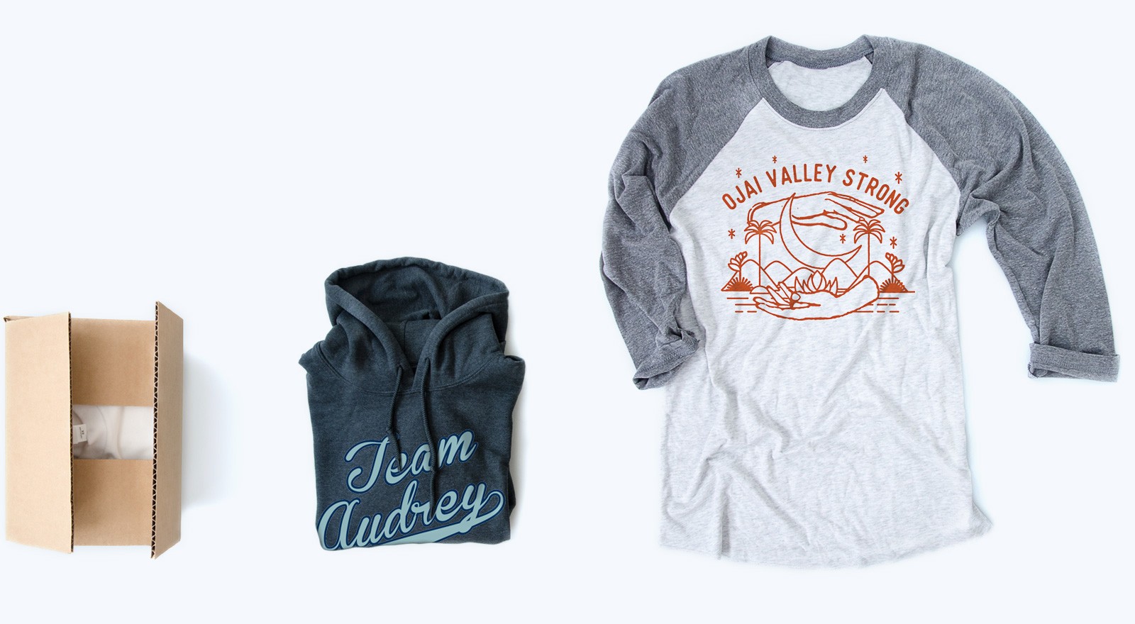 Cardboard box, folded Team Audrey hoodie, and Ojai Valley Strong raglan T-shirt on white background.