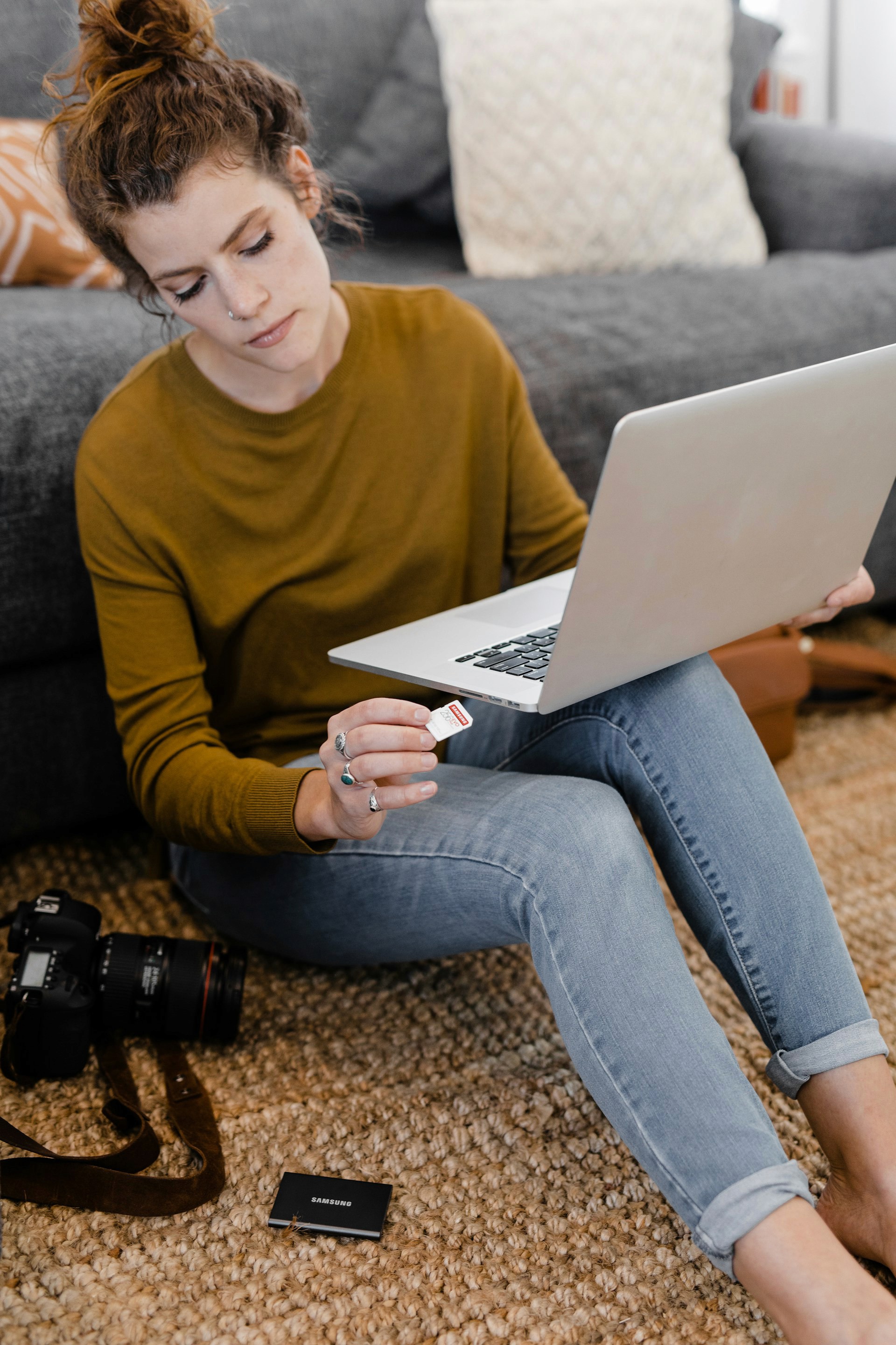 A woman in a mustard yellow top and blue jeans is sitting on a rug, holding a laptop and a small memory card, with a DSLR camera and lens beside her.