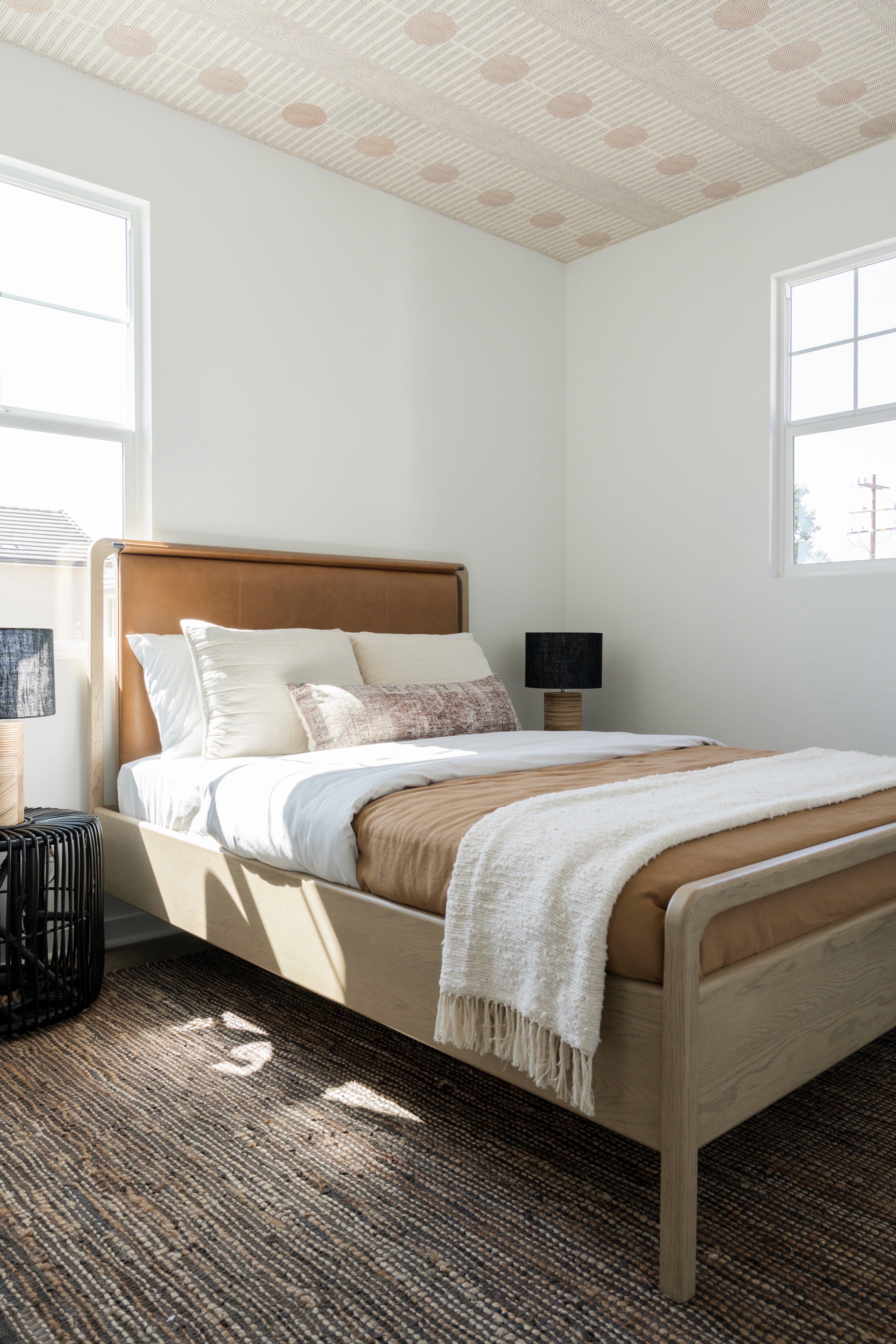 Bedroom with camel headboard and light wood frame, cream bedding and neutral accent pillows, black rope side table, and cream table lamps with black shades by Jubilee Interiors in Arcadia, California