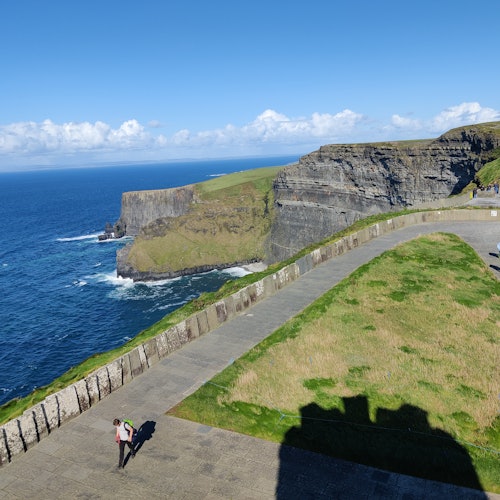 Човек стои на пътека близо до тревисти скали с изглед към синьо море под ясно небе.