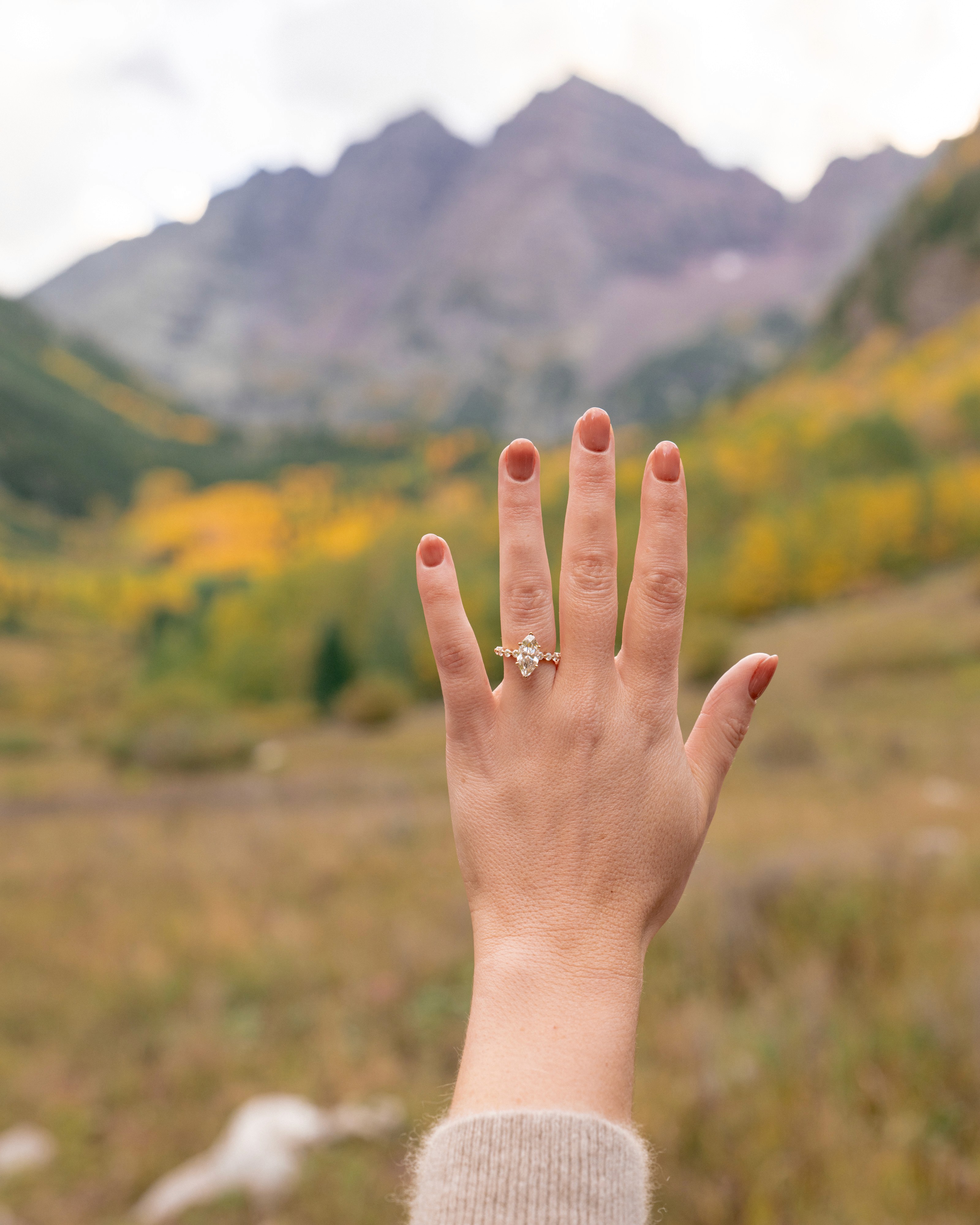Lexi holds up her left hand to show her engagement ring against a softly blurred mountain backdrop.