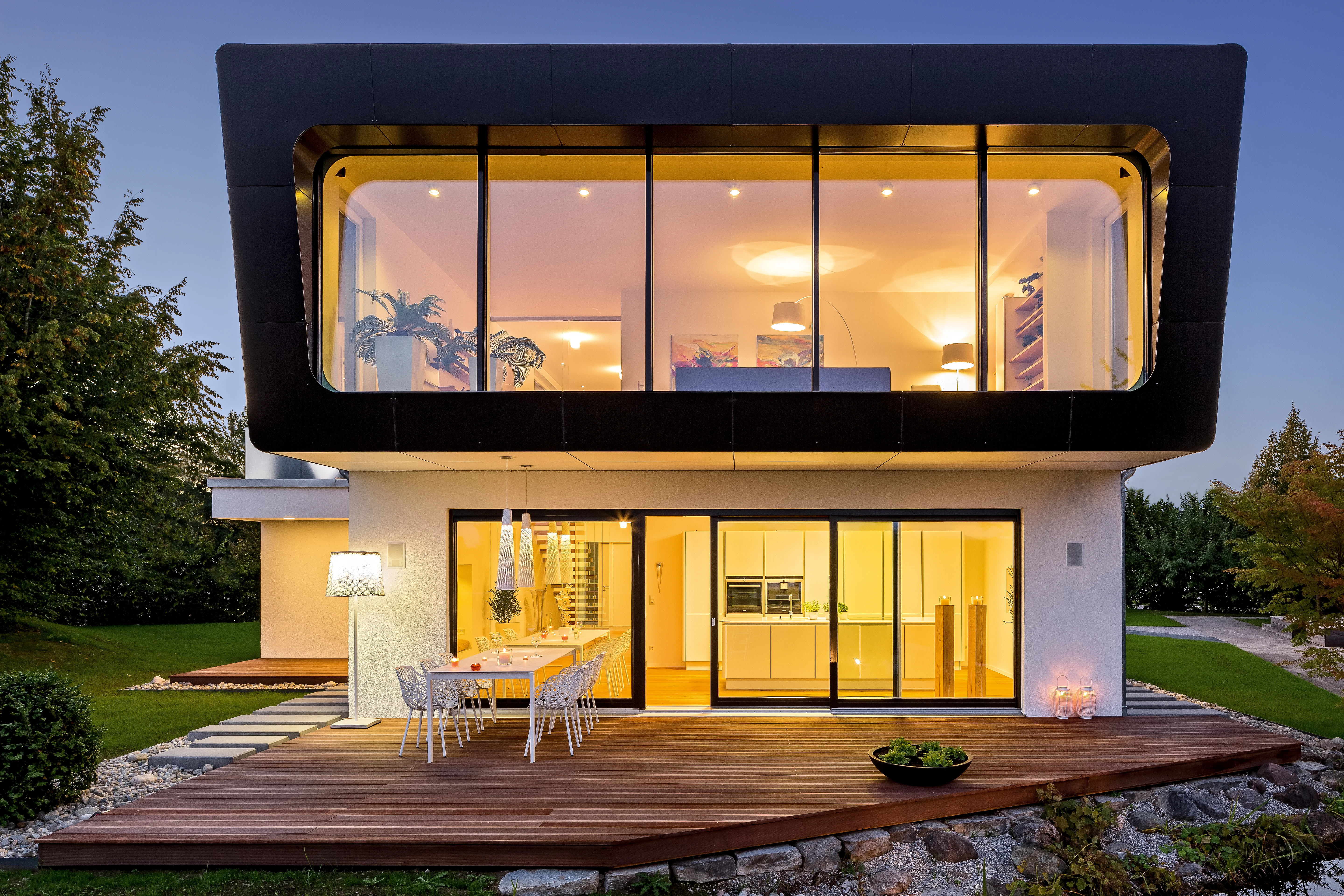 Modernes Wohnhaus mit großflächigen Fensterelementen, Holzterrasse mit Essgruppe und beleuchtetem Garten im Abendlicht.
