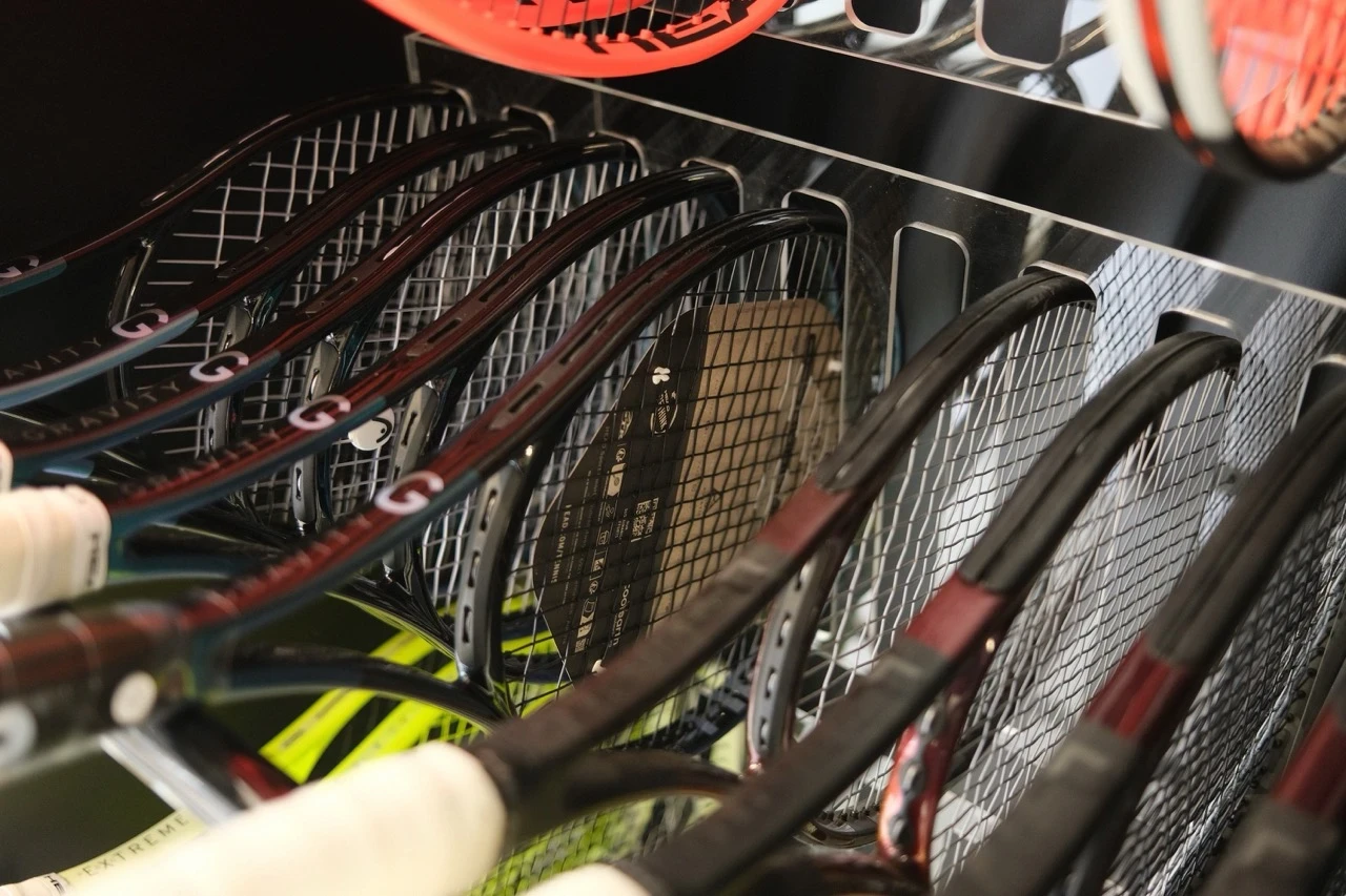 Eine Reihe von Tennisschlägern, hauptsächlich der HEAD Gravity Serie, ordentlich in einem Regal angeordnet. Die Schläger sind unbespannt, und ihre Rahmen schimmern im Licht. Im Hintergrund sind weitere Tennisschläger und reflektierende Oberflächen sichtbar.